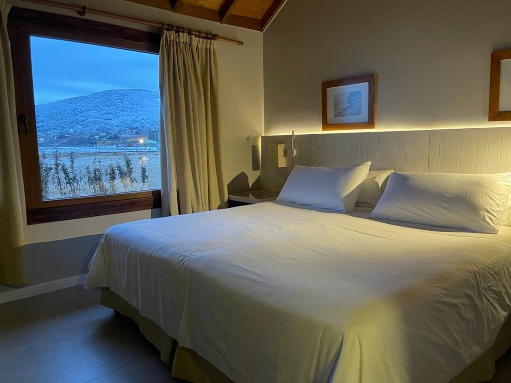 Un dormitorio con una cama grande, una mesita de noche, una lámpara de noche, una ventana con cortinas y un cuadro en la pared.
