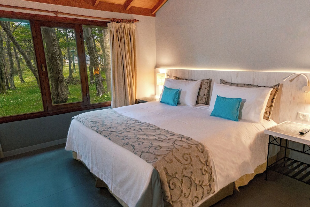 A bedroom with a large bed, two bedside lamps, a nightstand, and a window with curtains.
