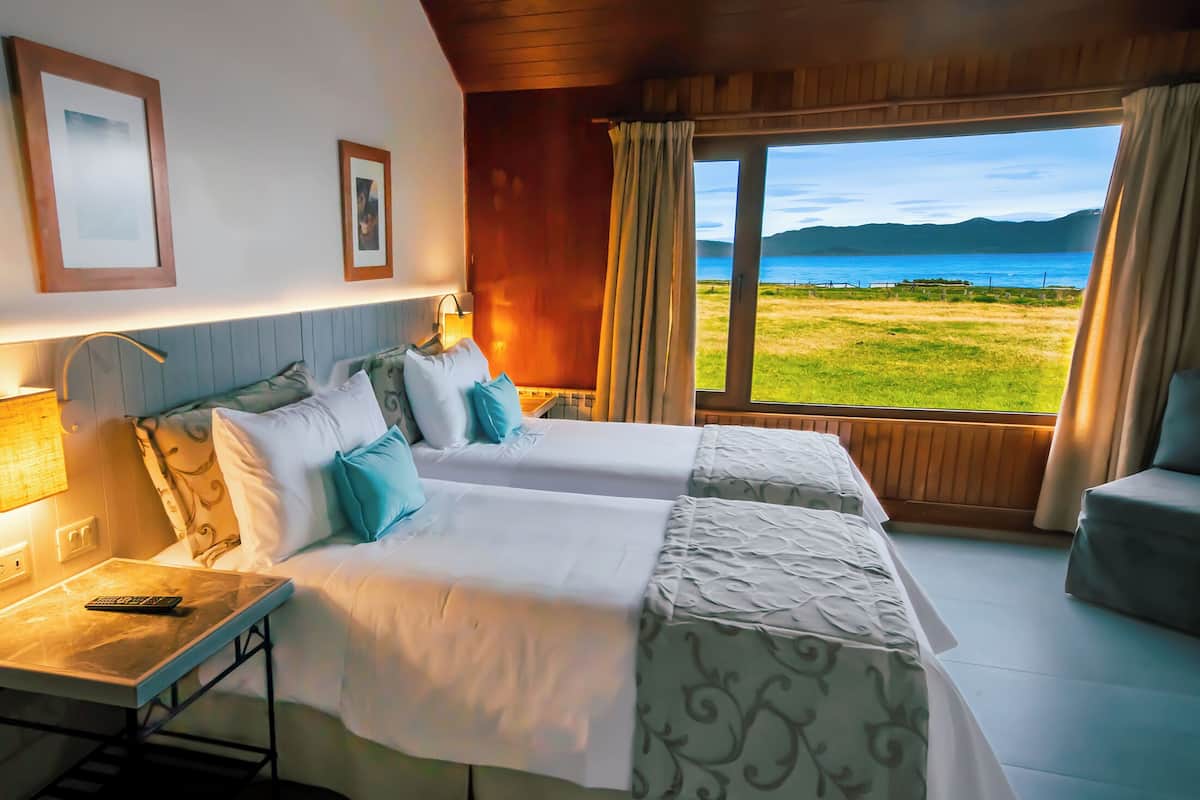 A hotel room with two beds, a desk, a lamp, and a view of the sea.