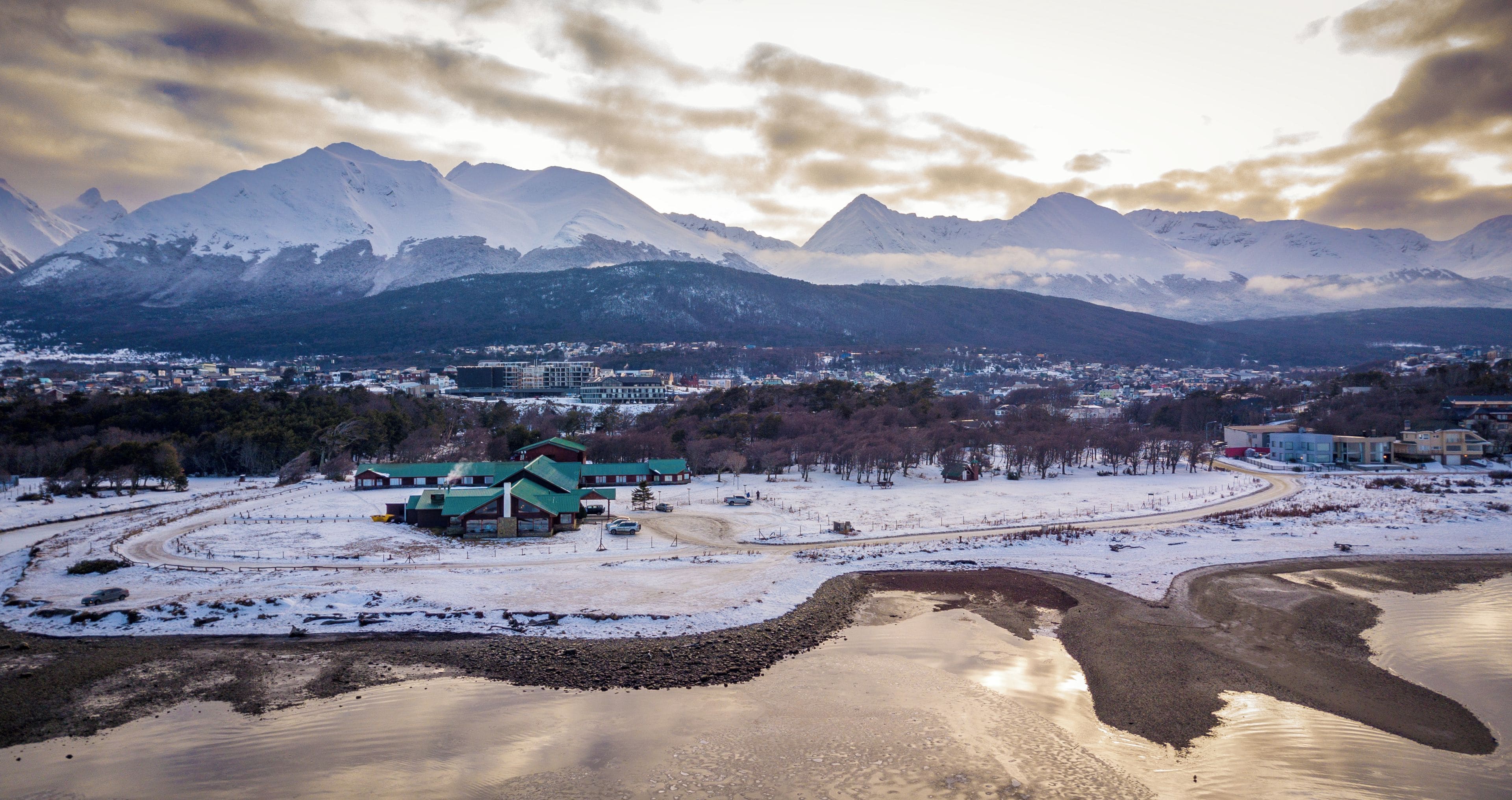 Paisaje nevado con un edificio de techo verde, rodeado de montañas y una pequeña masa de agua.