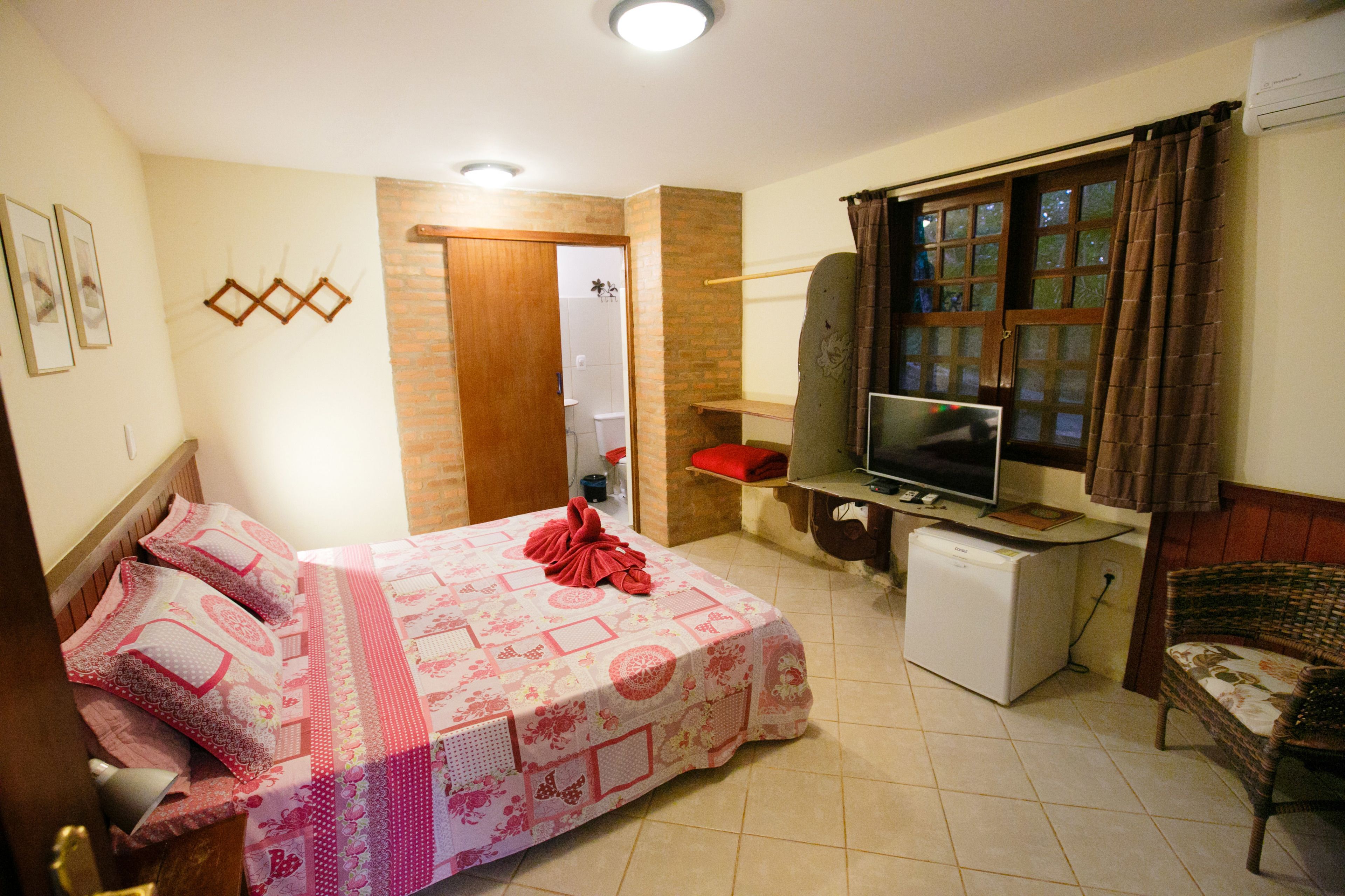 A bedroom with a bed, a television, a small refrigerator, and a wicker chair.