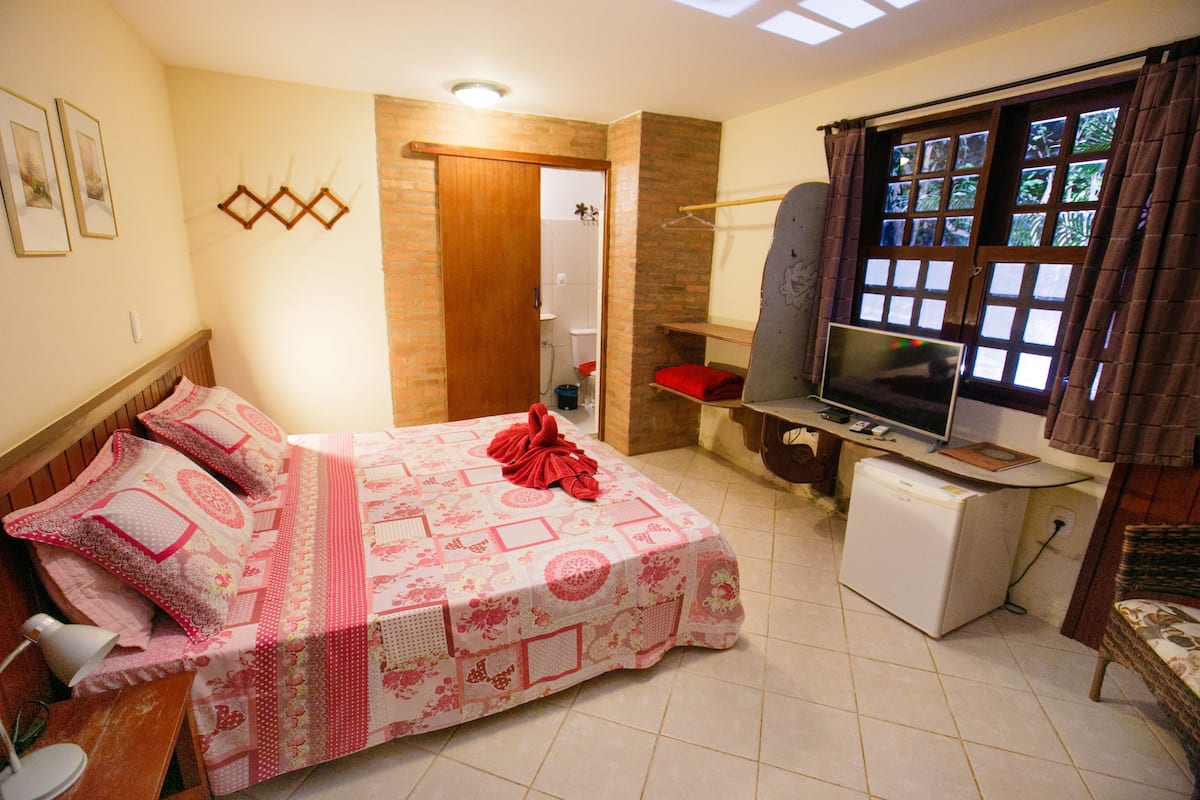 Quarto com cama, escrivaninha com televisão, mini-frigorífico e janela com cortinas.