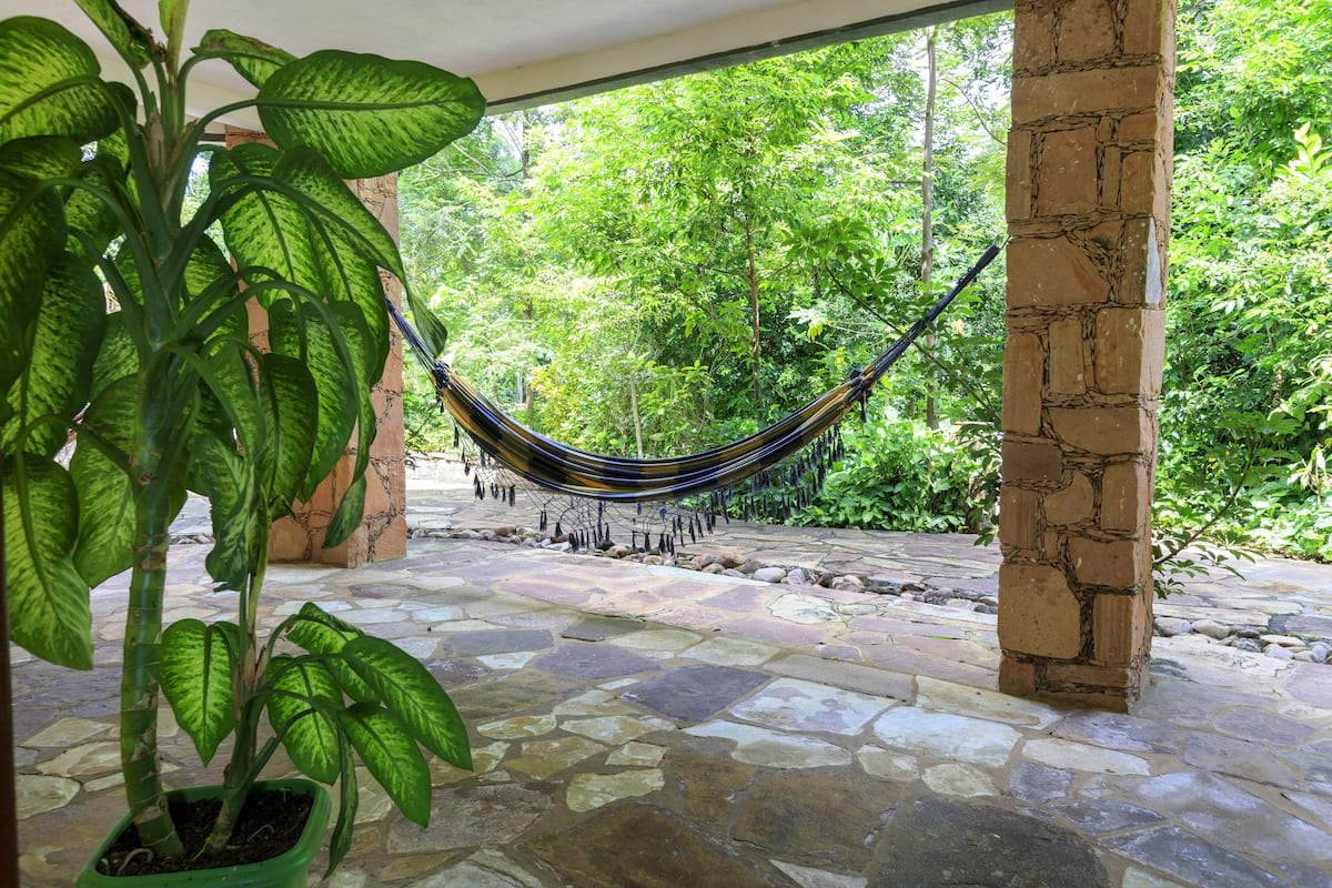 Uma rede sob uma área coberta com piso de pedra e um vaso de planta por perto.