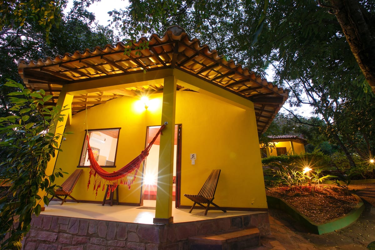 Um edifício amarelo com pérgola de madeira, duas cadeiras de madeira e uma rede.