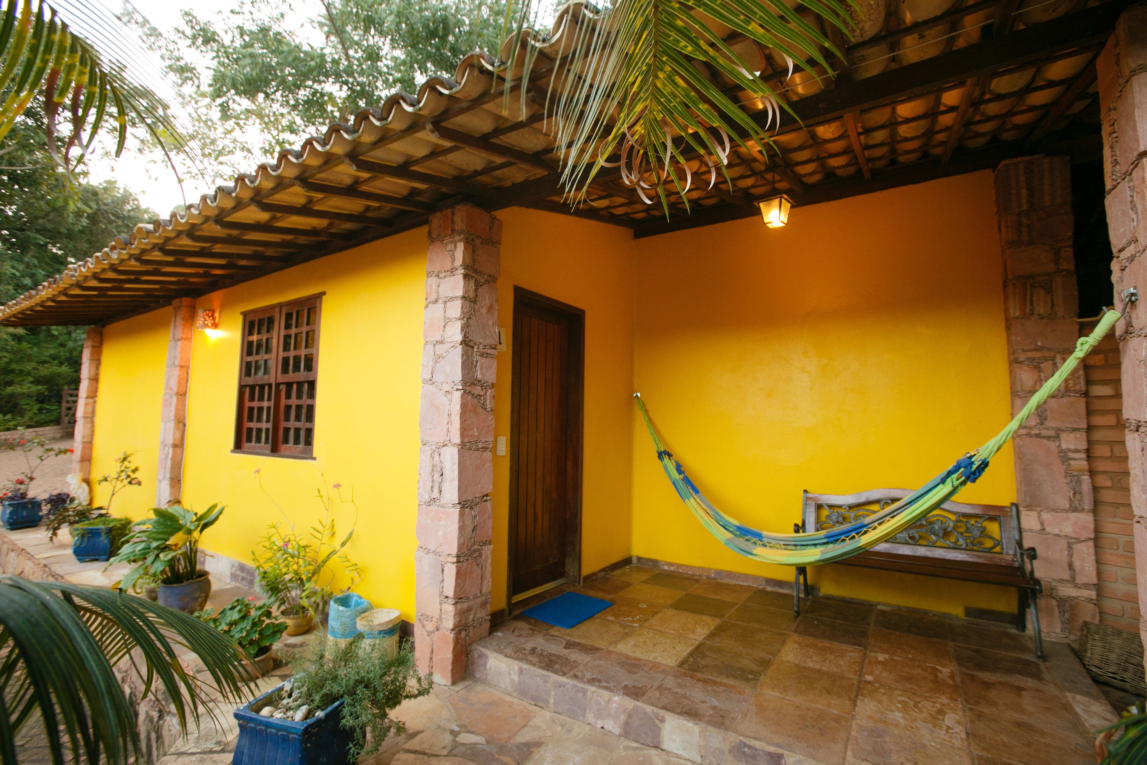 A yellow house with a tiled porch, a hammock, and potted plants.