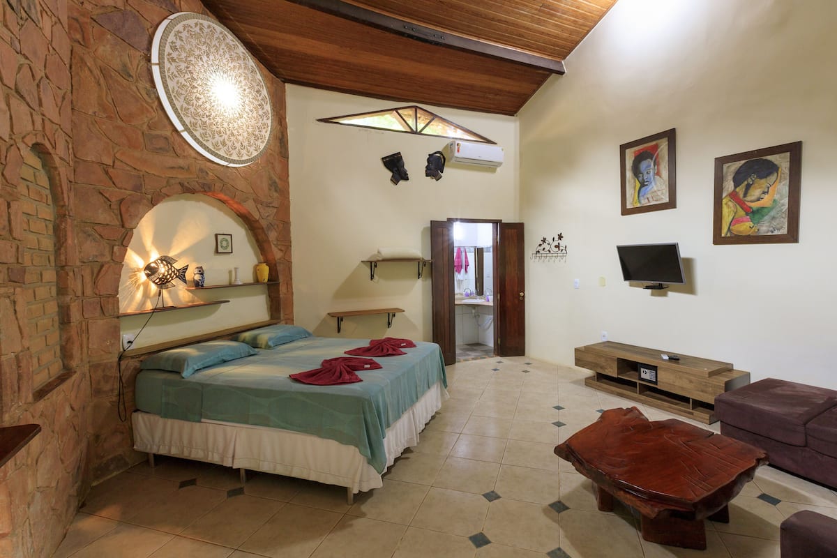 Um quarto com parede de pedra, uma cama com colcha verde e branca, teto de madeira, um banco de madeira, uma TV e obras de arte na parede.