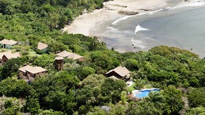 Een resort met meerdere bungalowachtige gebouwen, omgeven door weelderige tropische begroeiing, een zandstrand en een helderblauw zwembad.