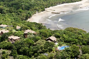 Resort com vários edifícios em estilo de bangalô, rodeado por vegetação tropical densa, uma praia de areia e uma piscina azul cristalina.