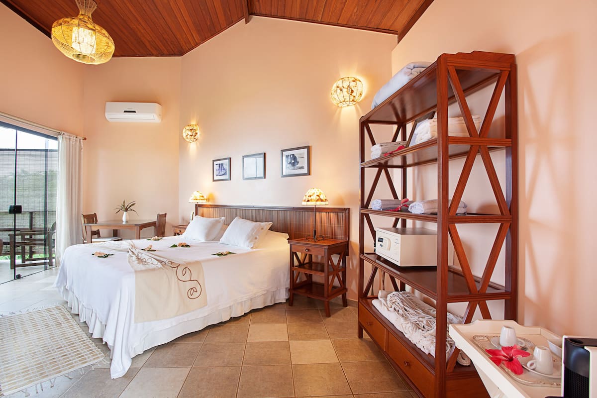 Une chambre d’hôtel comprenant un lit, une table de chevet, une lampe et une étagère en bois où sont posées des serviettes et des articles de toilette.