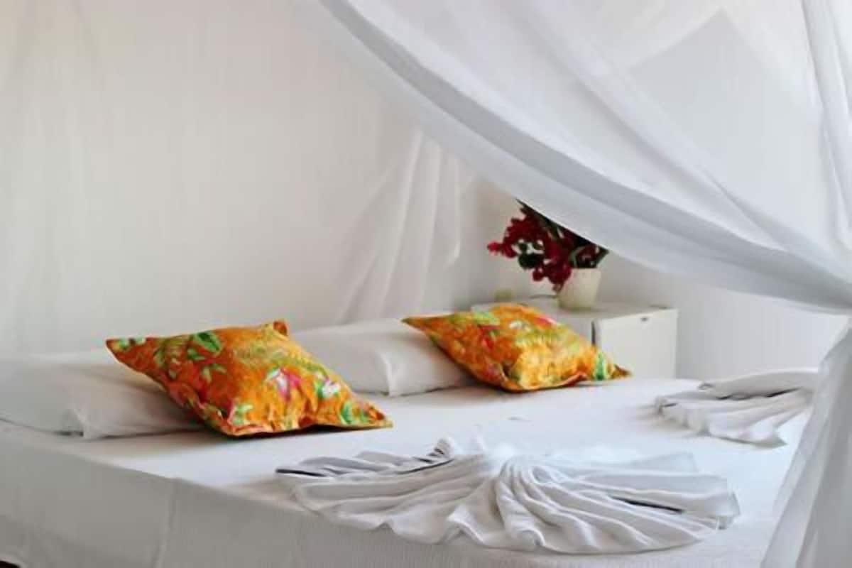 A bed with white bedding and colorful pillows, a white bedside table with a vase of flowers, and a sheer white canopy over the bed.