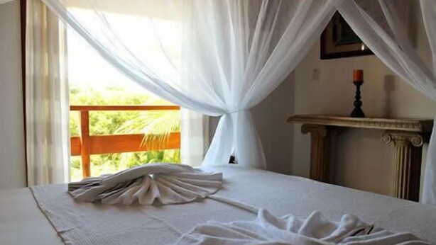 A hotel room with a bed, a window with sheer curtains, a wooden headboard, and a small table with a candle. - Casa Grande & Senzala (Porto Seguro)