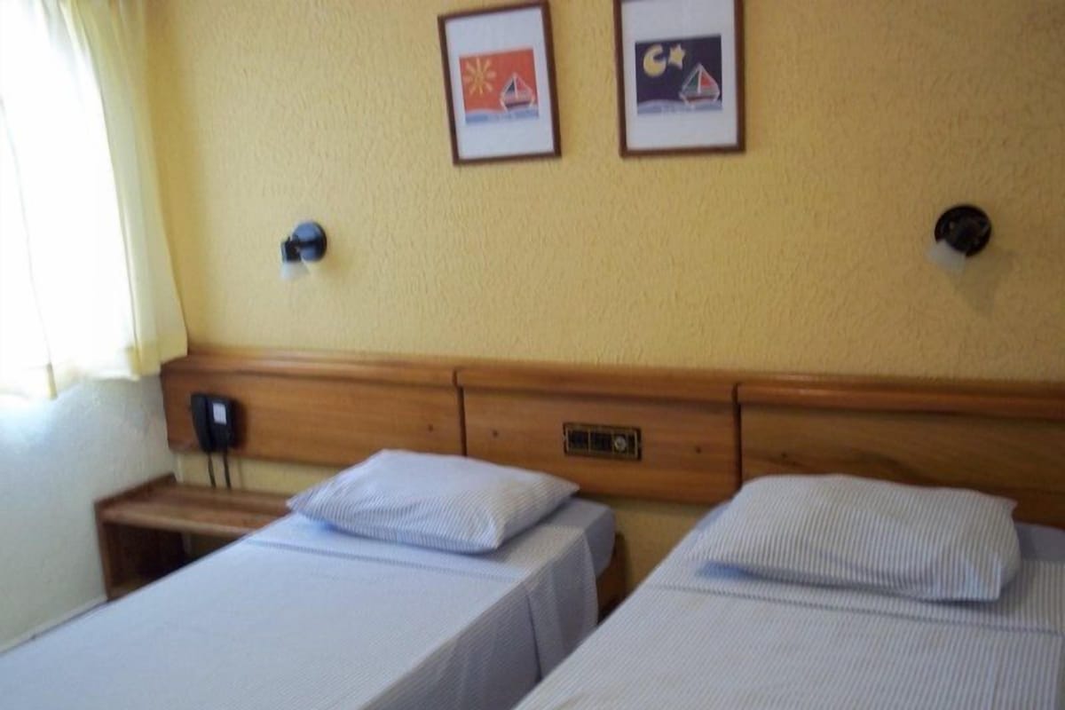 A hotel room with two single beds, a wooden headboard, a bedside table with a lamp, and a wall-mounted phone.