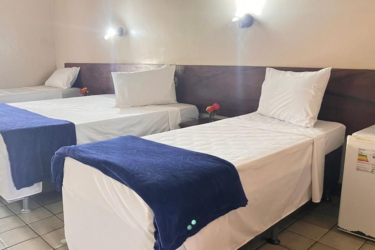 Habitación de hotel con tres camas individuales, cada una con colchón blanco y colcha azul. Entre las camas hay una mesita con un florero.