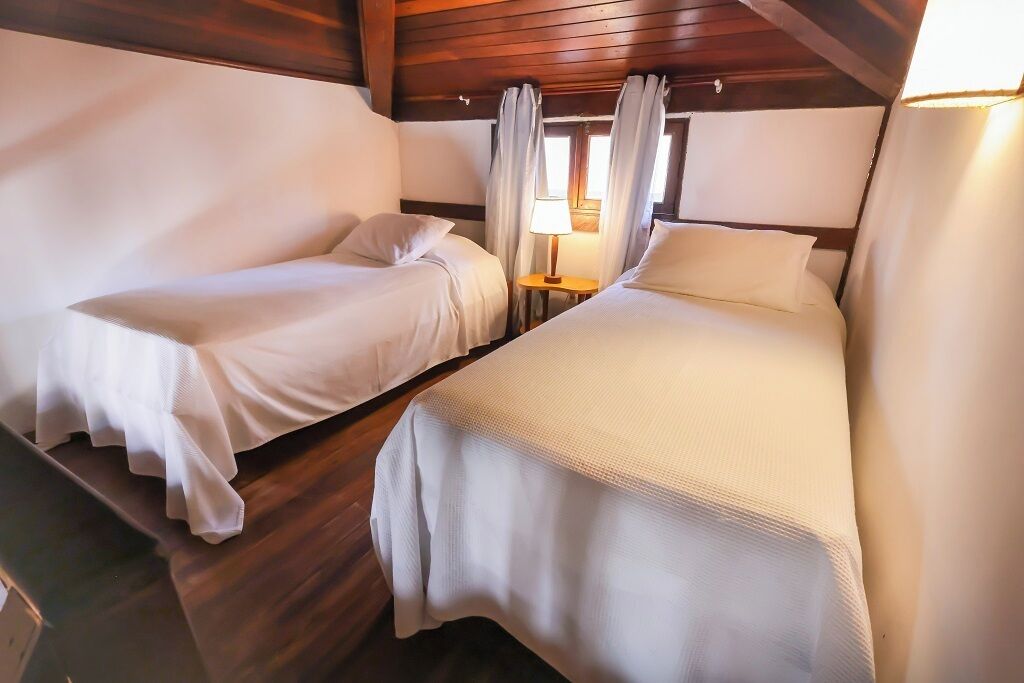 Une cabine équipée de deux lits simples, d’une petite table avec une lampe, de murs en bois et d’une fenêtre avec des rideaux.