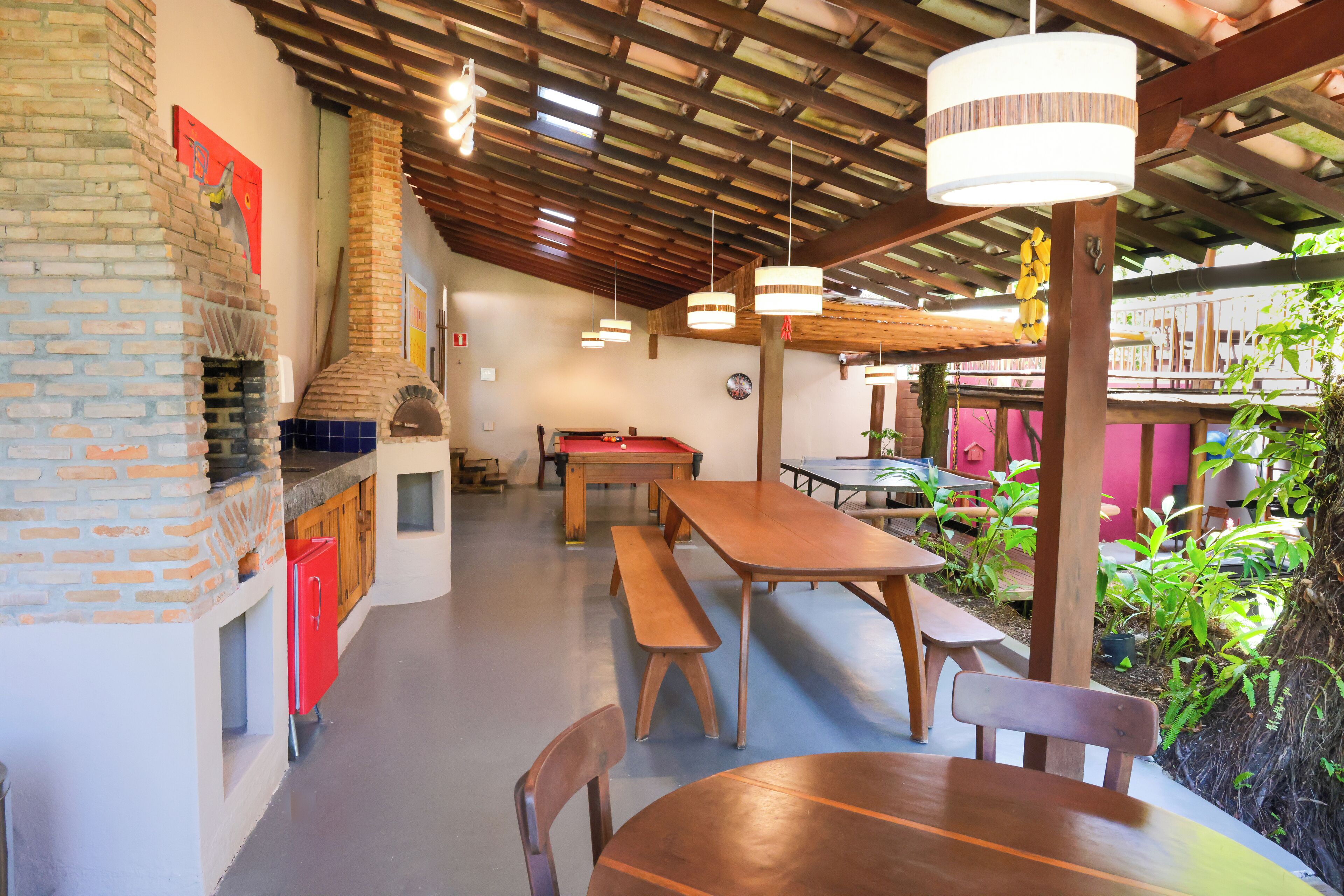 A spacious indoor dining area with a brick fireplace, wooden tables, benches, and hanging pendant lights.