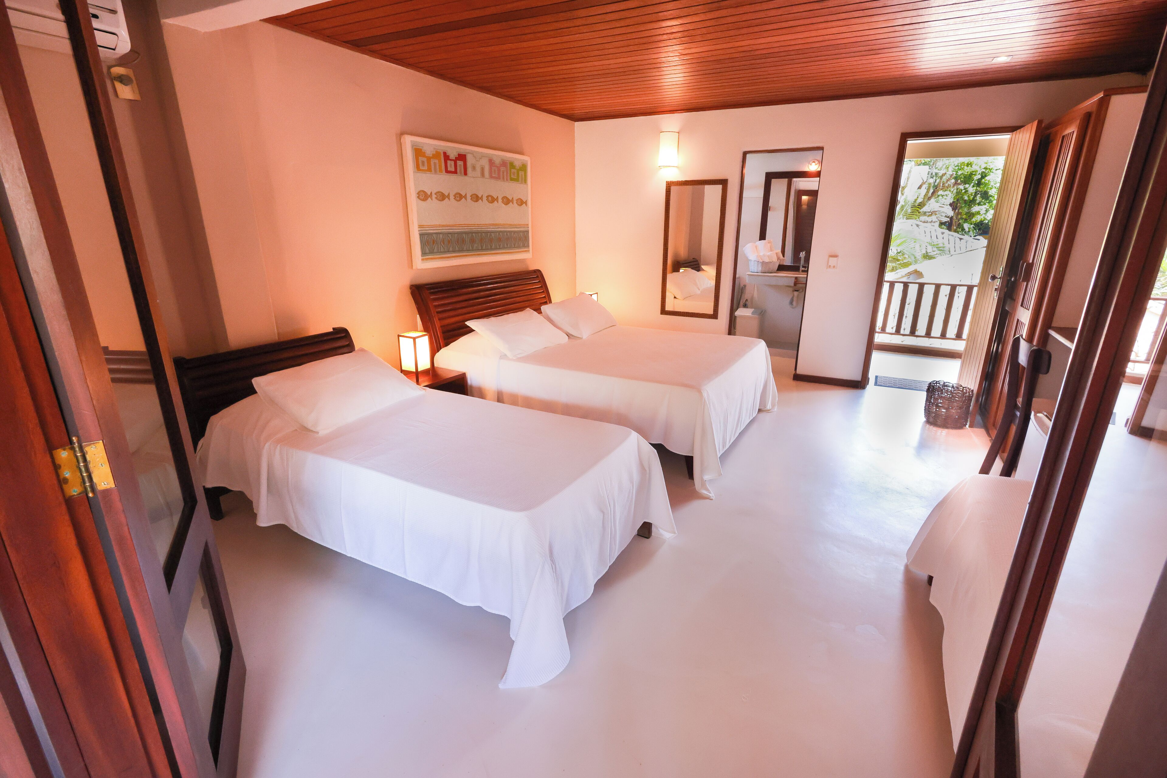 A hotel room with two beds, a wooden ceiling, a large mirror, and a balcony with a view of trees.