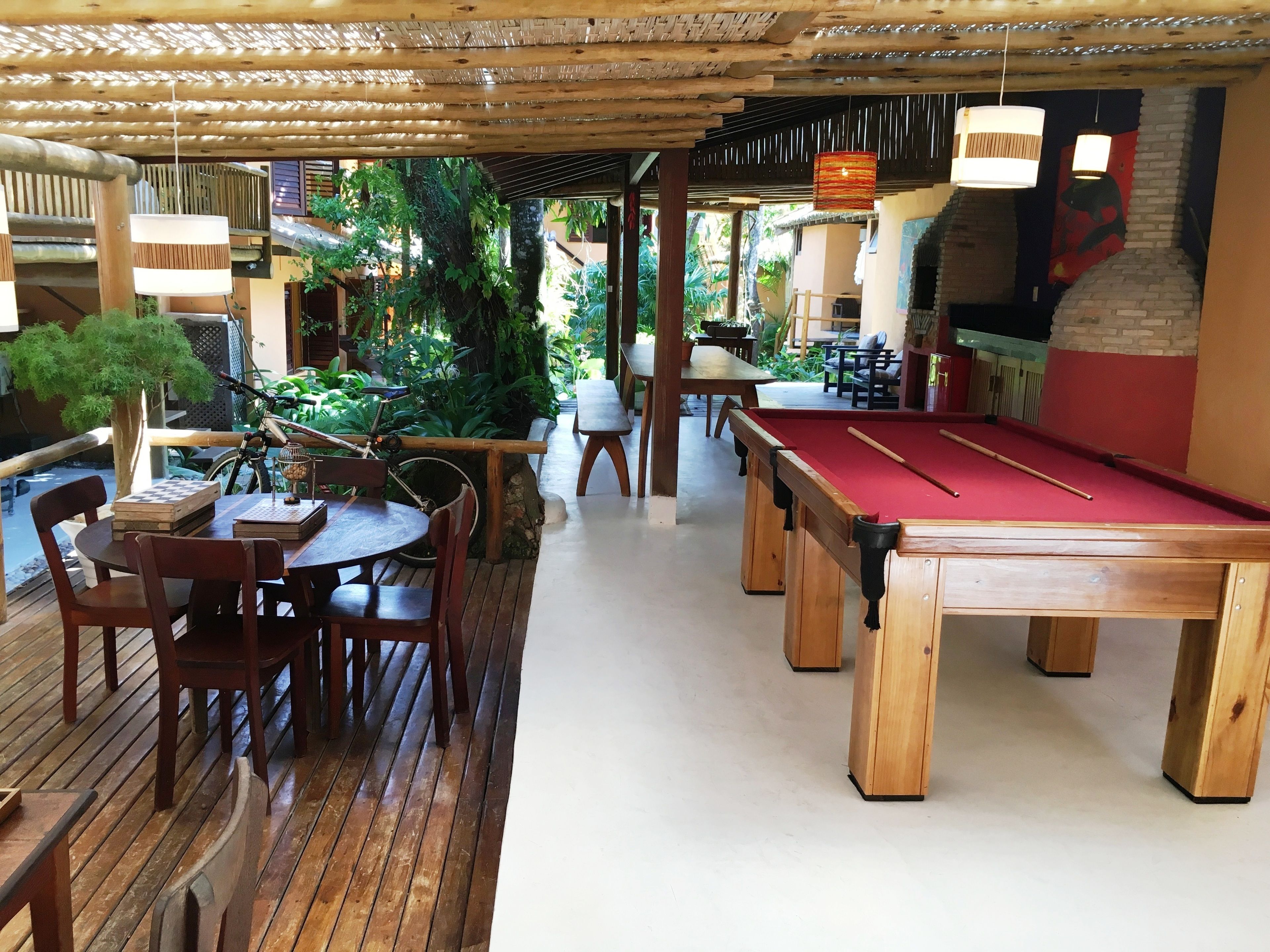 An indoor recreational area with a pool table, wooden tables, and chairs, surrounded by lush greenery and a wooden deck.