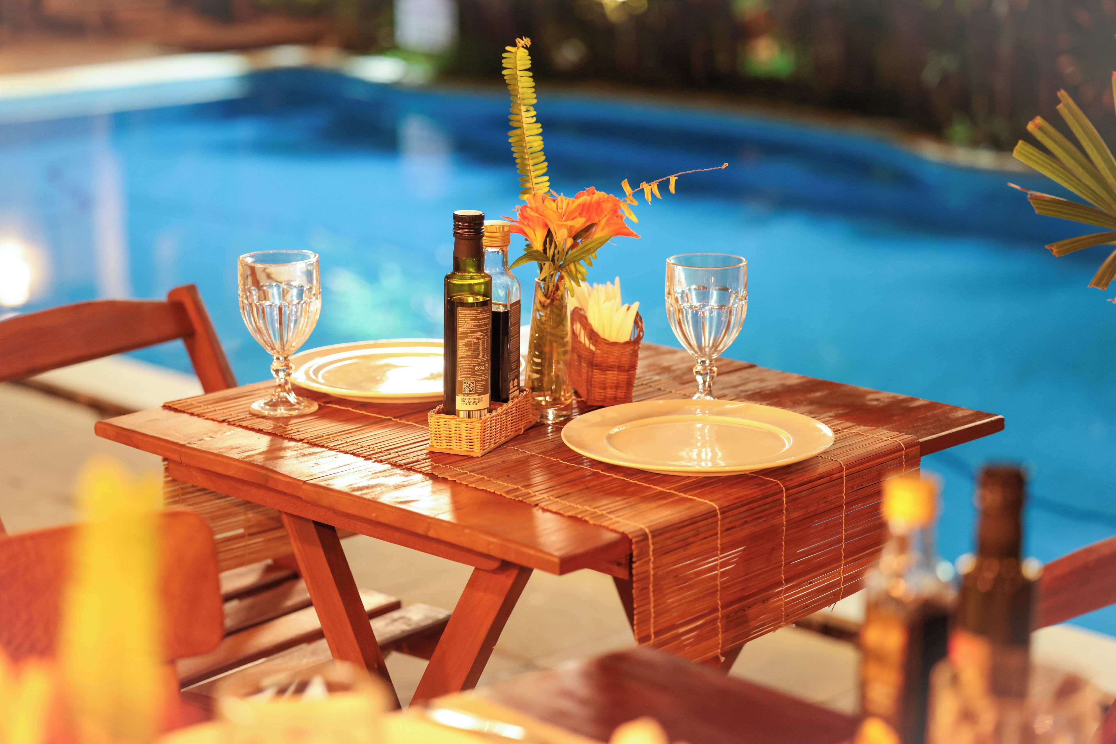 Een houten tafel gedekt voor twee personen, met een fles wijn, een glas en een vaas bloemen bij een zwembad.