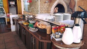 Montaje de bufé con diversos alimentos, una máquina de café y un horno de ladrillo al fondo.