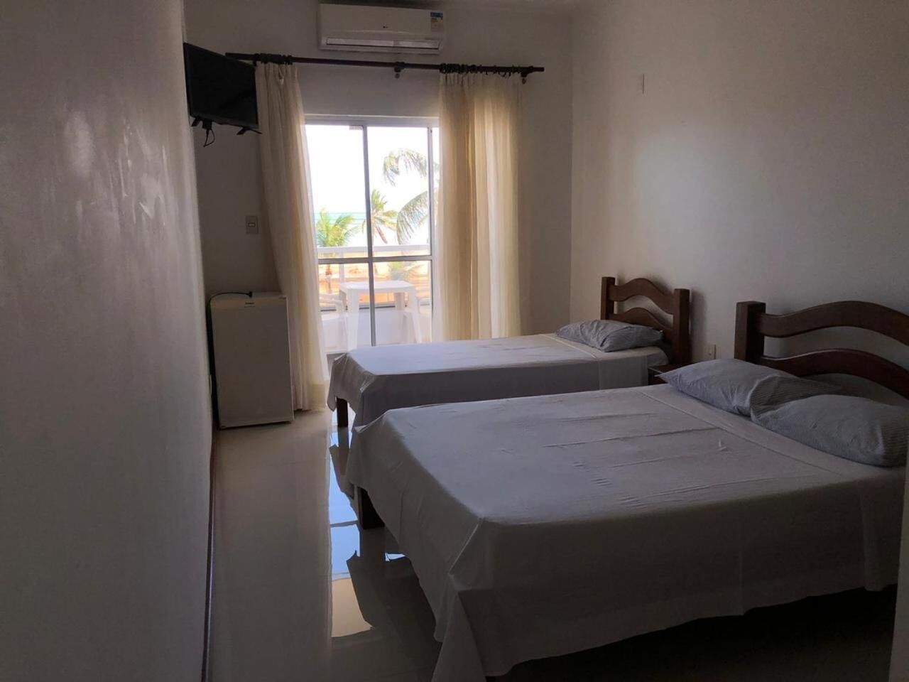 A room with two beds, a window with curtains, and a view of palm trees.