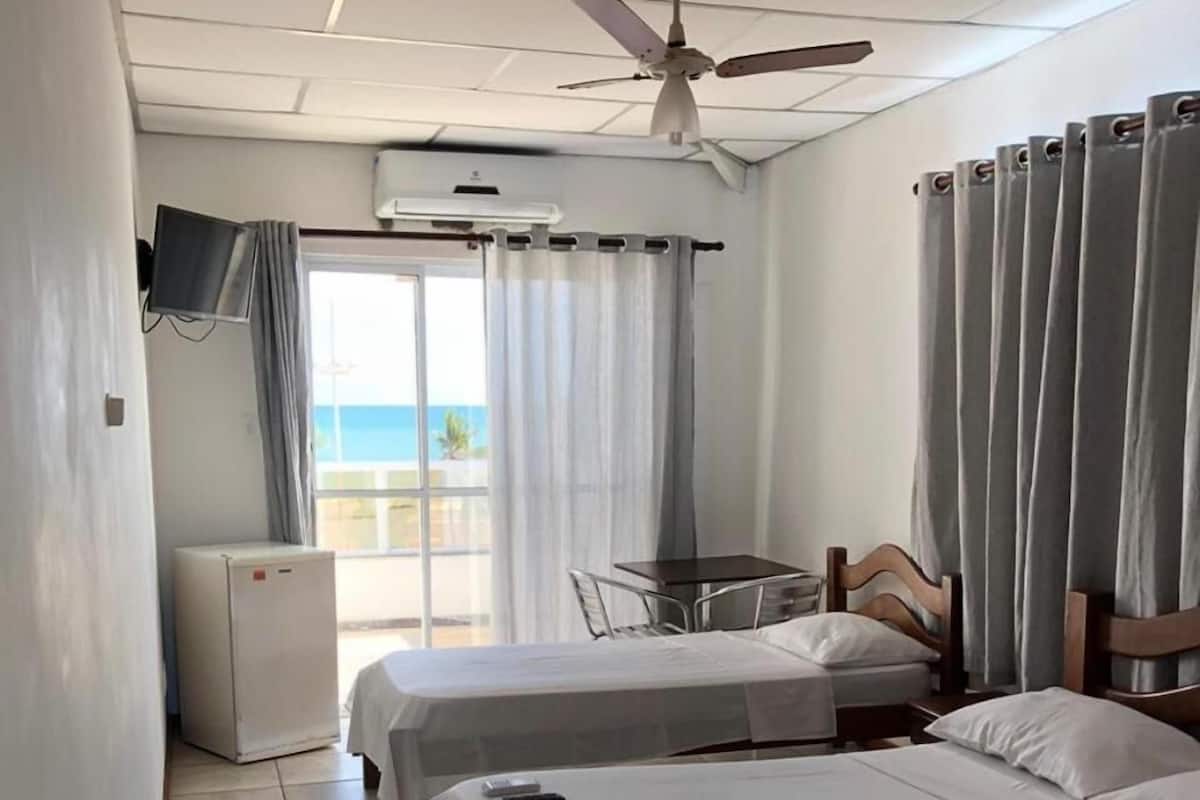 A hotel room with two beds, a small table, a TV, a ceiling fan, and a view of the ocean.