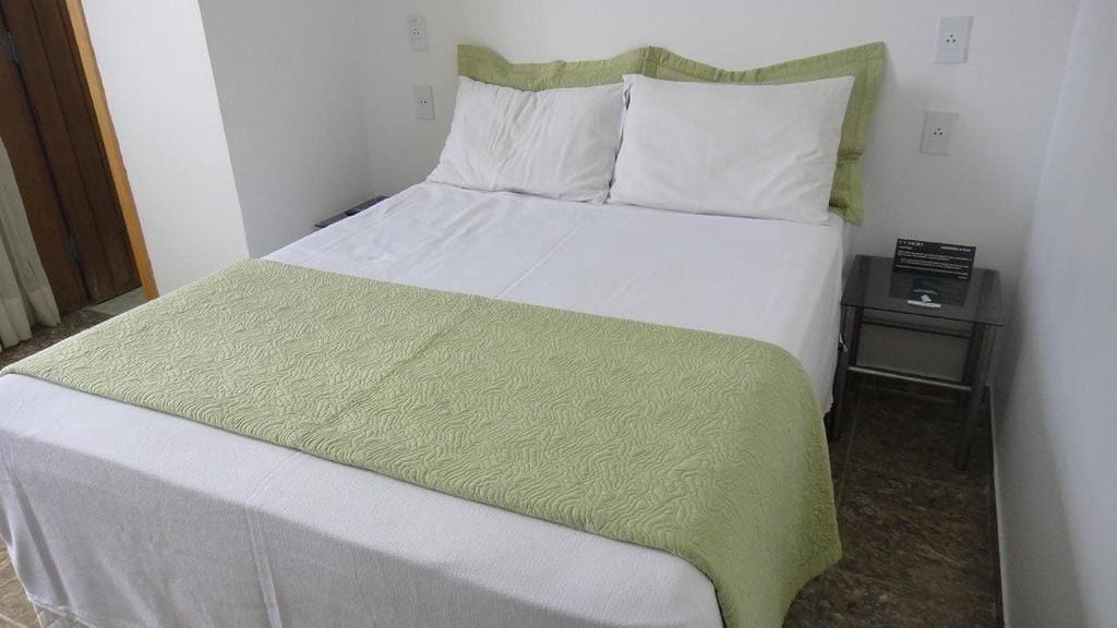 A neatly made bed with a green and white bedspread, a small bedside table with a lamp, and a wall-mounted power outlet.