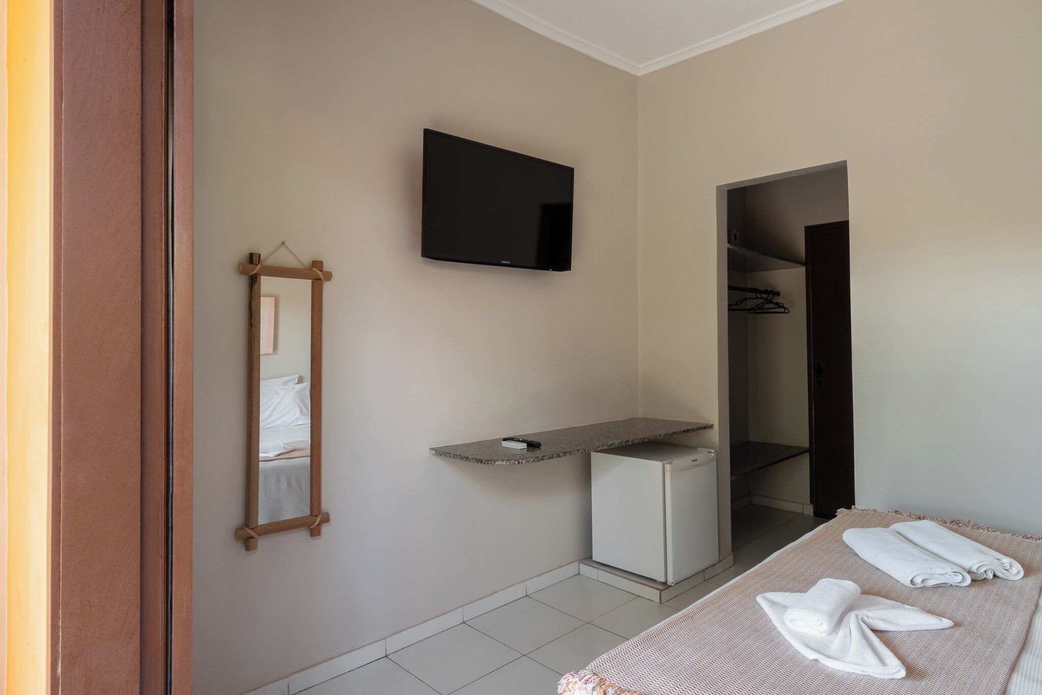 A hotel room with a flat-screen TV mounted on the wall, a small refrigerator, a wooden-framed mirror, and a bed with neatly folded towels.