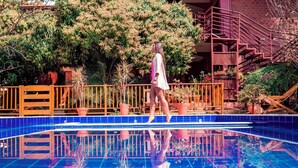 Une personne se promène sur une piscine dont le fond est recouvert de carreaux bleus, et dont la surface reflète l’environnement. Des plantes en pot et une clôture en bois l’entourent.