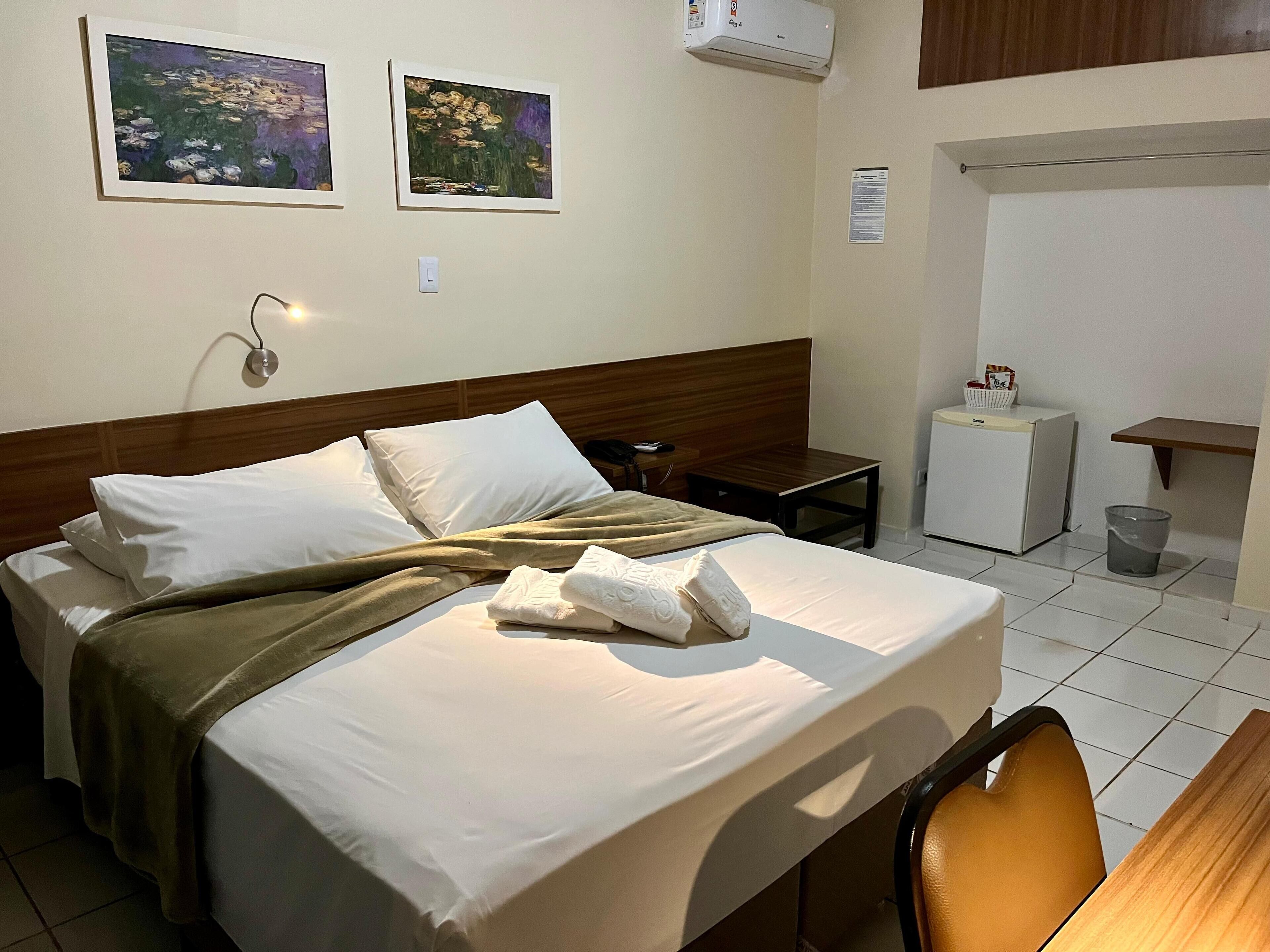 A neatly made bed with white linens and a brown throw blanket, a small bedside table with a lamp, a refrigerator, and a trash can.