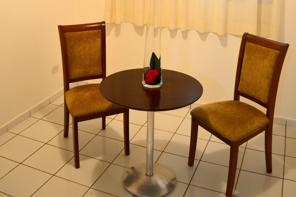 A room with a round table, two chairs, a window with curtains, and a painting on the wall.