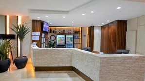 Zona de recepción con mostrador, televisor, refrigerador y plantas en macetas.