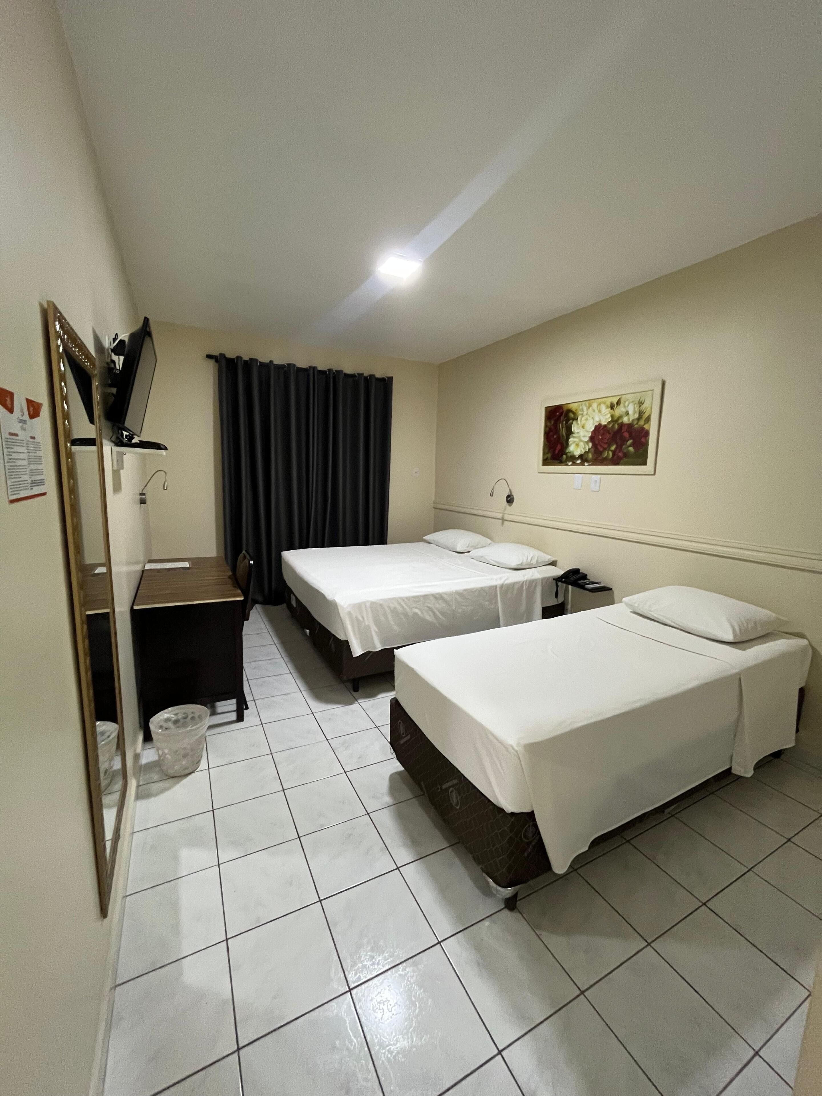 A hotel room with two single beds, a desk, a chair, a mirror, a trash bin, and a painting on the wall.