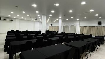 Un salón de conferencias con filas de mesas y sillas, iluminación en el techo y un equipo de aire acondicionado.