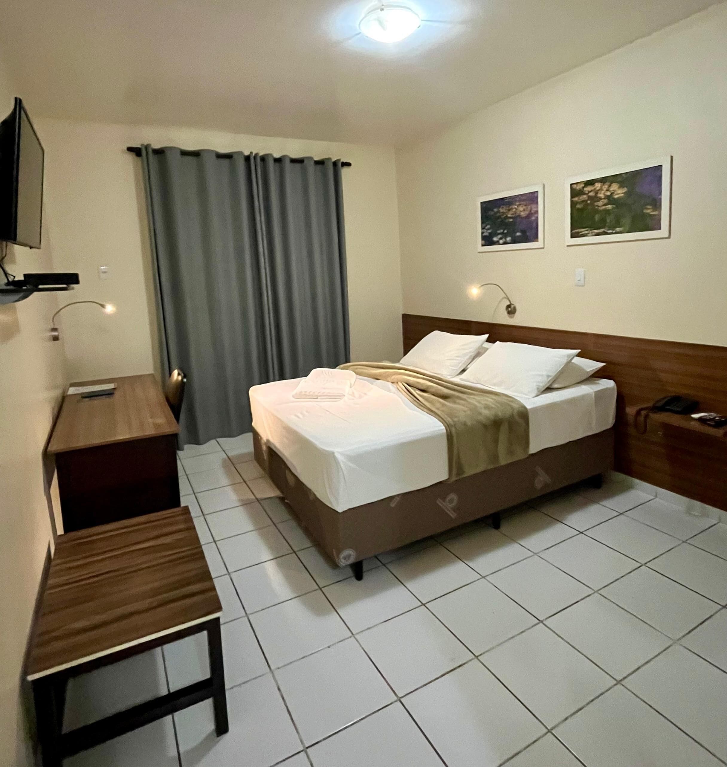 Quarto de hotel com cama, escrivaninha, cadeira, televisão e janela com cortinas.