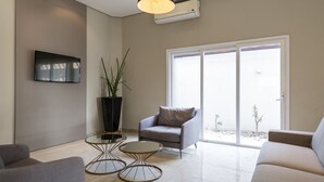 A modern living room with a grey sofa, a small armchair, and a coffee table. There is a large window with a view of a garden. - Hotel Tainá - Aeroporto Cuiabá (Varzea Grande)