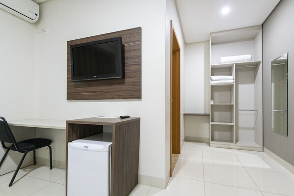 A compact hotel room with a flat-screen TV mounted on a wooden panel, a small desk with a chair, a compact refrigerator, and a walk-in closet with shelves.
