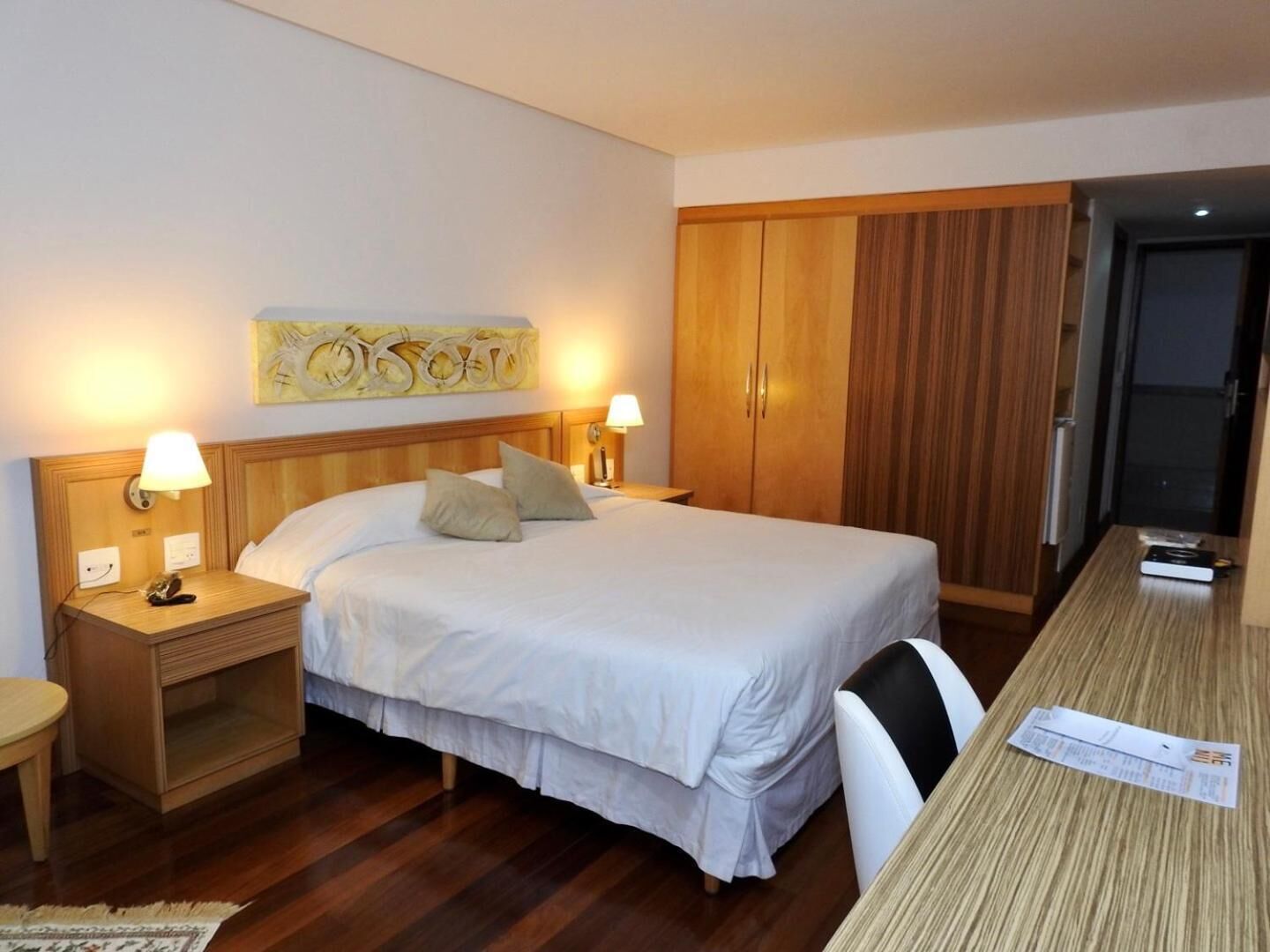 A hotel room with a bed, bedside tables, a desk, a chair, and a wardrobe.