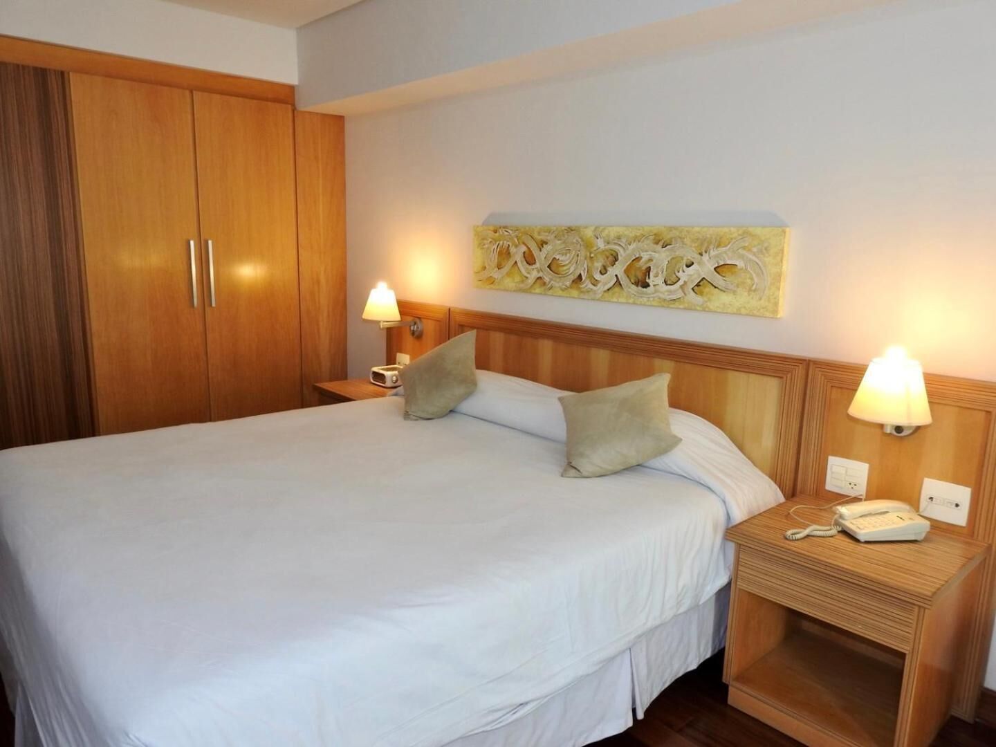 Quarto de hotel com uma cama grande, duas mesas de cabeceira, um guarda-roupa de madeira e um objeto decorativo na parede.