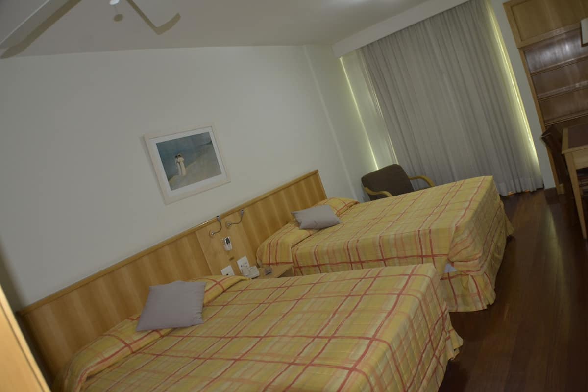 Quarto de hotel com duas camas, uma cadeira, uma escrivaninha e um quadro na parede.