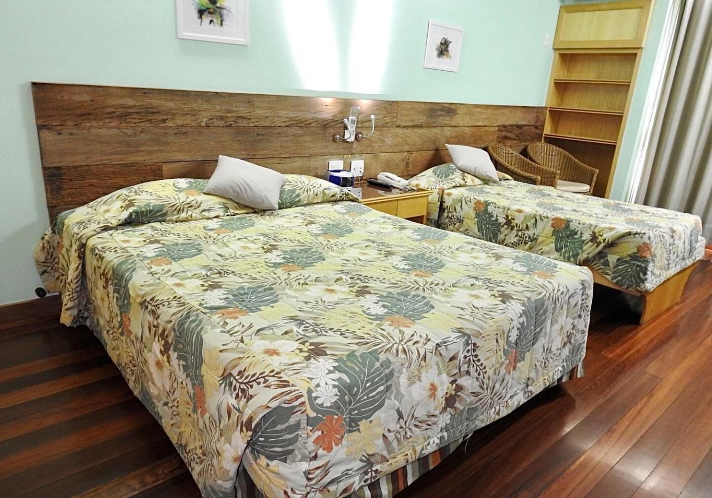 A hotel room with two beds, wooden headboard, floral bedspreads, a small bedside table with a lamp, and a shelf with baskets.