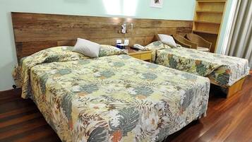 A hotel room with two beds, wooden headboard, floral bedspreads, a small bedside table with a lamp, and a shelf with baskets.