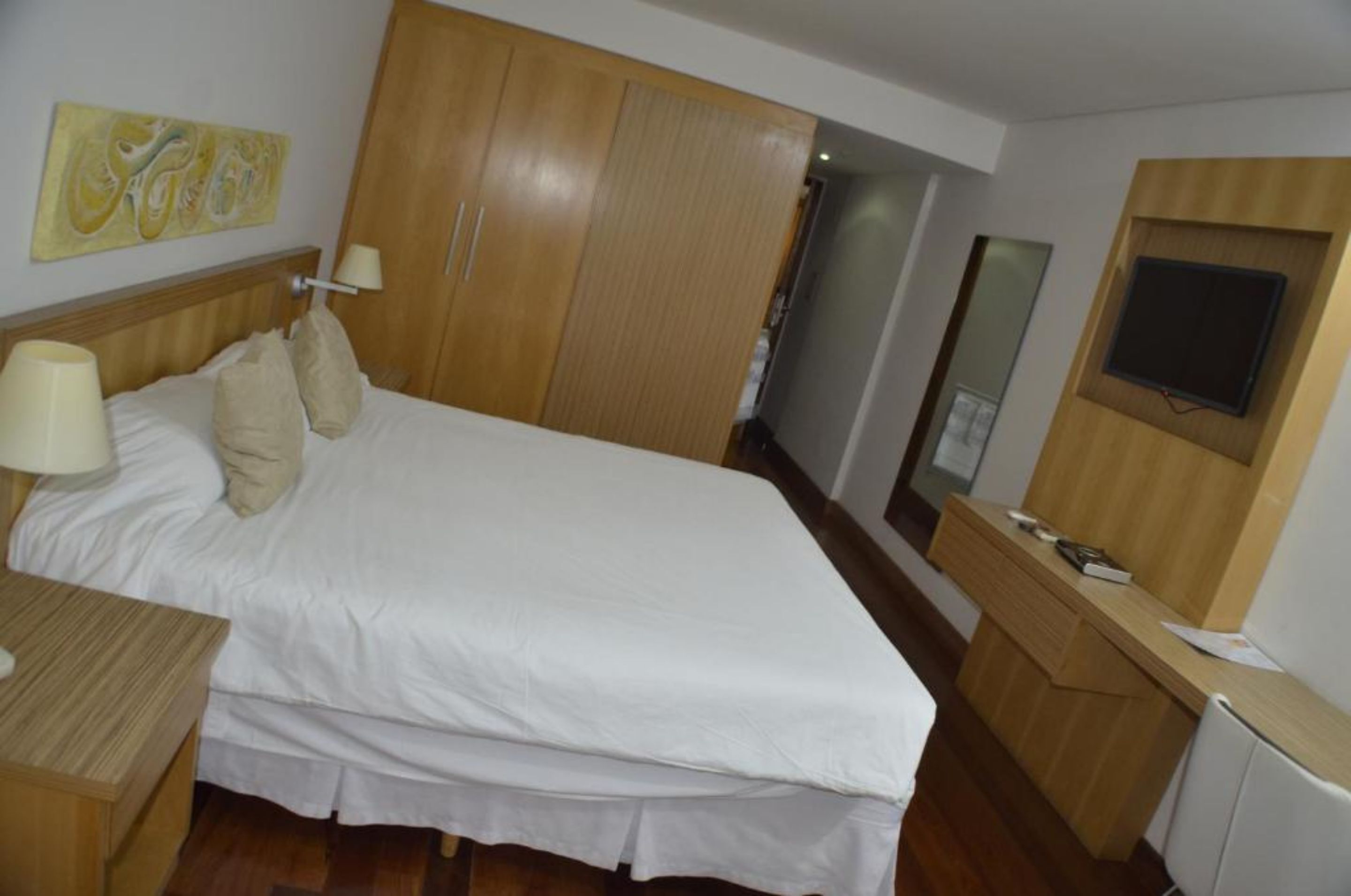 A hotel room with a bed, bedside table, lamp, wardrobe, and a TV mounted on the wall.