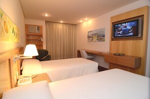 Quarto de hotel com duas camas, uma escrivaninha, televisão, abajur e um quadro na parede.