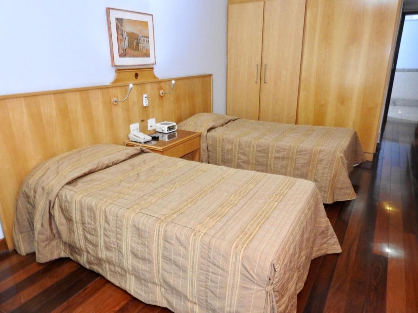 A hotel room with two beds, wooden headboard, a bedside table with a phone and radio, and a framed picture on the wall.