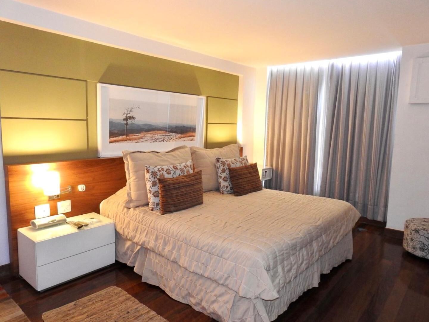 A bedroom with a large bed, a nightstand, a lamp, a framed picture on the wall, and a window with curtains.