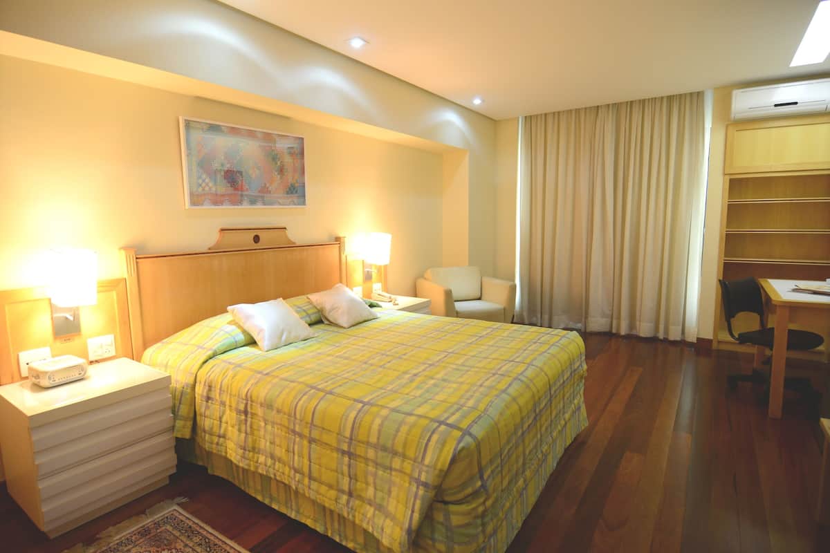Quarto de hotel com cama, mesas de cabeceira, escrivaninha, cadeira e ar condicionado.