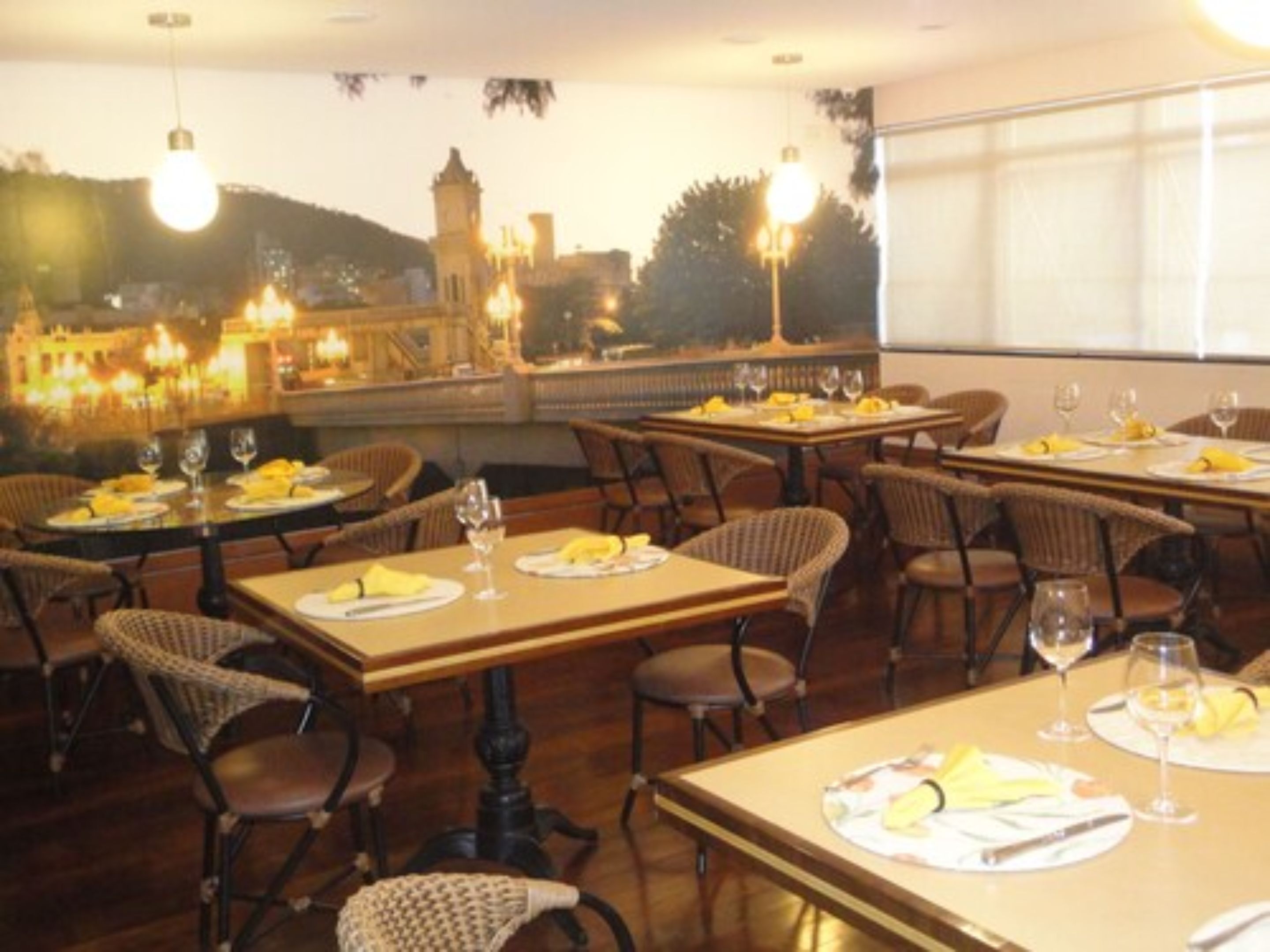 A restaurant with tables set for dining, featuring wicker chairs and a view of a cityscape with a tower.