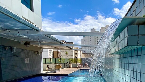 A swimming pool with clear blue water, a water fountain feature, and a view of the surrounding buildings. - Constantino Hotel (Juiz de Fora)