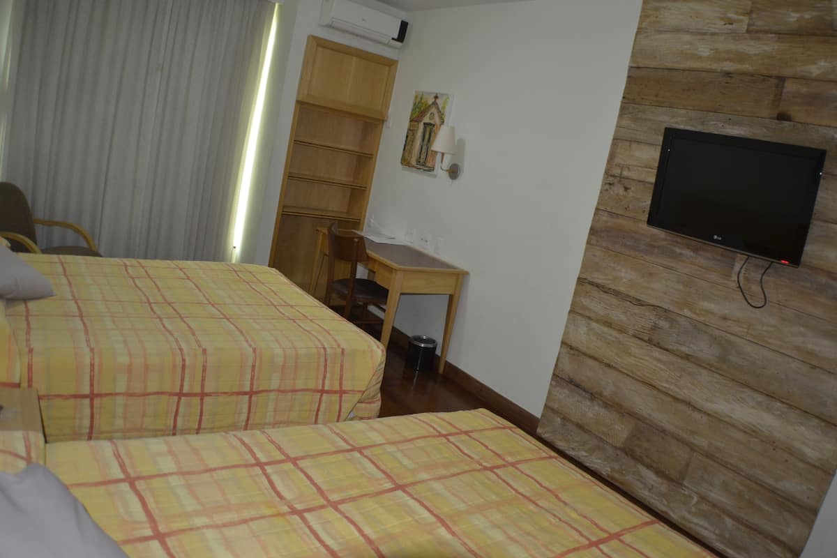 Quarto de hotel com duas camas, uma escrivaninha, uma cadeira, uma televisão e uma janela com cortinas.