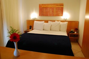 Una habitación de hotel con una cama, mesitas de noche, un jarrón con una flor y un cuadro en la pared.